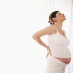 Pregnant woman holding back in front of white curtain